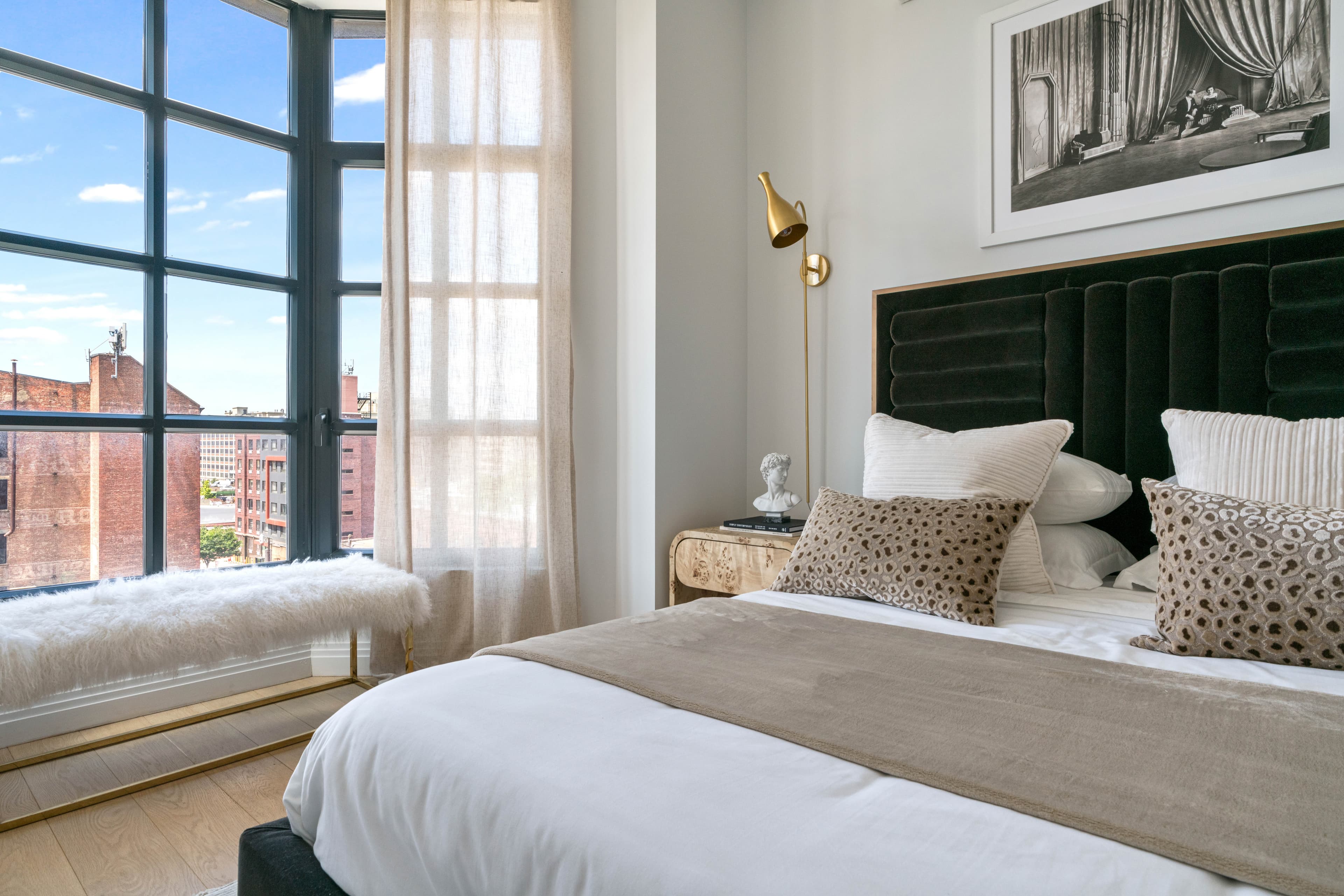 A staged bedroom at 875 Fourth Avenue with a channel-tufted headboard and a corner window over low-rise Brooklyn rooftops