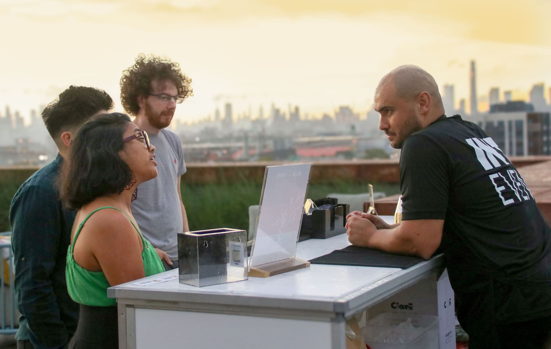 Guests gathered at the drinks table in golden-hour light with the Manhattan skyline behind them on the rooftop at 875 Fourth Avenue